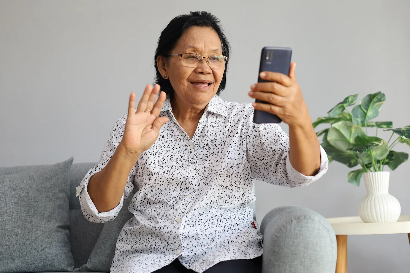 An older woman smiling and waving during a video call with family from her living room