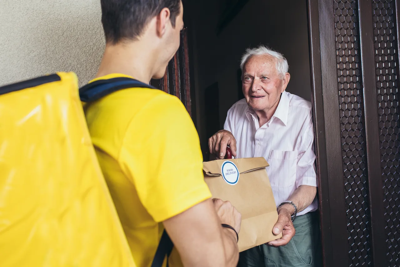 A delivery driver handing a bag of groceries to a smiling older man at his front door