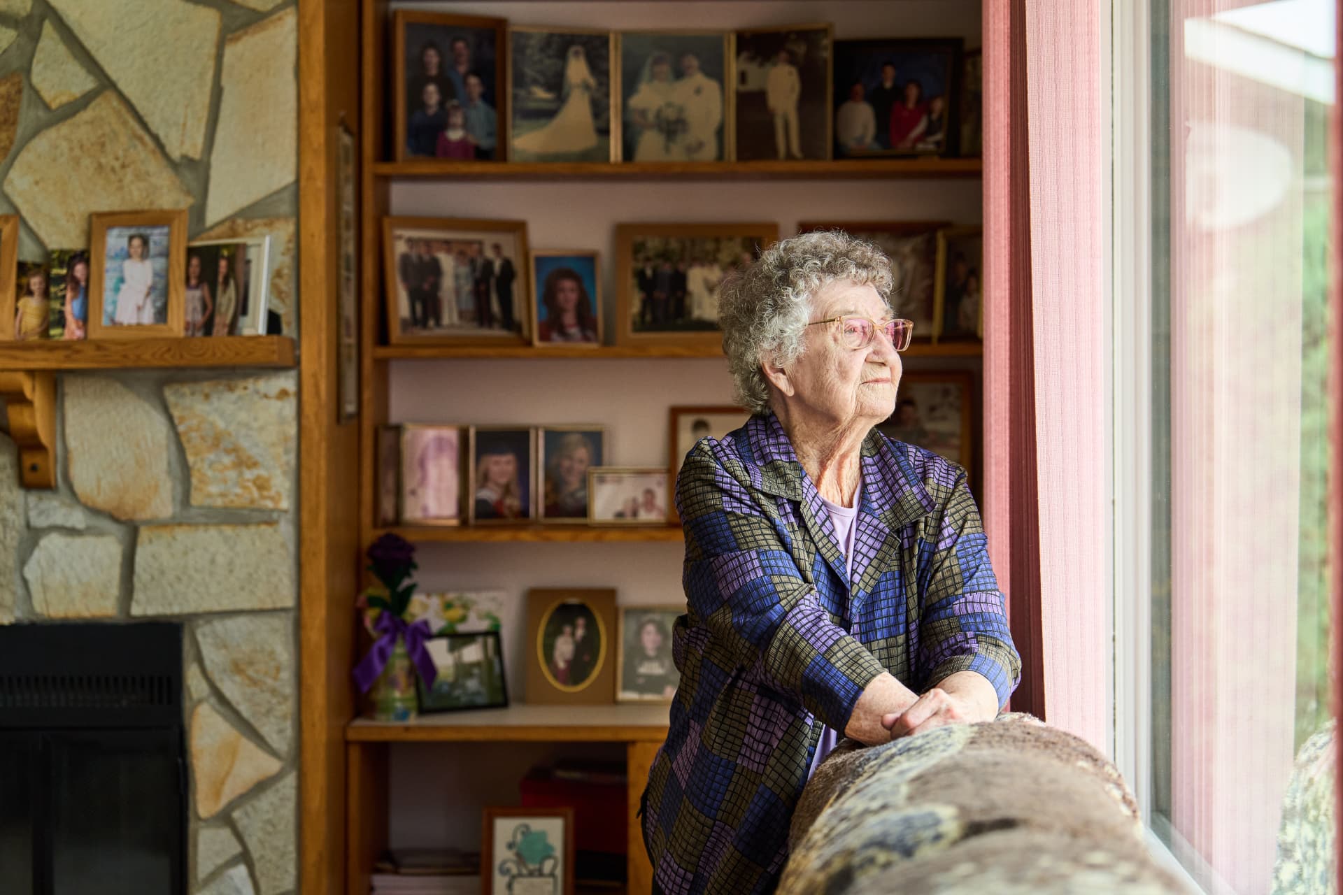 An older woman standing by a sunlit window in her living room, surrounded by framed family photos